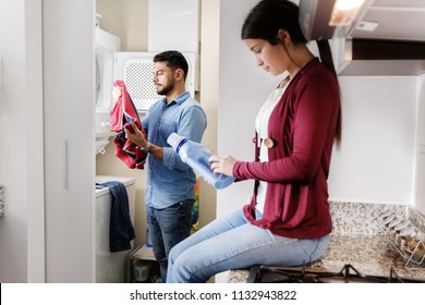 Young Couple Doing Housework And Chores. Man Putting Clothes Inside Washing Machine And Girlfriend Sitting On Kitchen Counter, Reading Label On Cleansing Box.