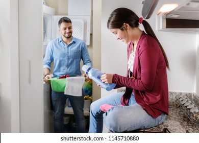 Young Couple Doing Housework And Chores. Man Putting Clothes Inside Washing Machine And Girlfriend Sitting On Kitchen Counter, Reading Label On Cleansing Box.