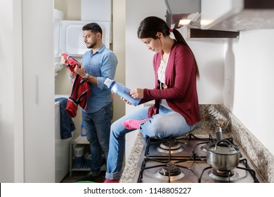 Young Couple Doing Housework And Chores. Man Putting Clothes Inside Washing Machine And Girlfriend Sitting On Kitchen Counter, Reading Label On Cleansing Box.