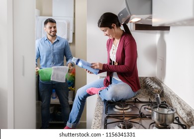 Young Couple Doing Housework And Chores. Man Putting Clothes Inside Washing Machine And Girlfriend Sitting On Kitchen Counter, Reading Label On Cleansing Box.