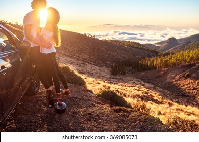 Young couple dressed alike in white t-shirt and hat embracing near the car on the roadside on the sunset.