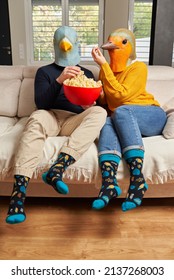 A Young Couple Dressed In Bird Masks Eat Popcorn On The Sofa At Home While Watching Series On Streaming Platforms