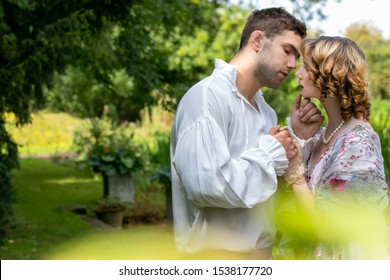 Young couple dressed in vintage costume standing on embankment by river and under trees, kissing.