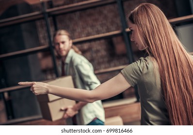 Young Couple Is Drifting Apart After Breakup, Girl Is Pointing To The Entrance, Man Is Carrying His Stuff In Cardboard Boxes