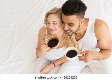 Young Couple Drink Coffee Sitting In Bed, Happy Smile Young Hispanic Man And Woman Top Angle View Lovers Hold Cups Bedroom