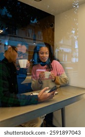 Young couple drinking coffee and using a smartphone, seen through a window, in a warm and cozy cafe