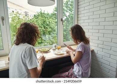 Young Couple Eating Fried Eggs During Breakfast At Table Of Windowsill. Concept Of Domestic Lifestyle. Idea Of Relationship And Spending Time Together. Morning Time. Modern Studio Apartment