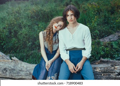 Young Couple Of Elves In Love Siting On Tree In Magical Forest Outdoor On Nature. Fairy Tale Love, Relationship And Magik People Concept. Man And Woman Looking At Camera