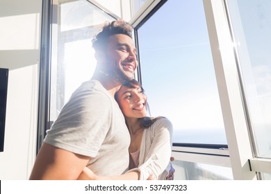 Young Couple Embrace Modern Apartment Big Panoramic Window Sea View, Mix Race Man And Woman Morning Home Interior