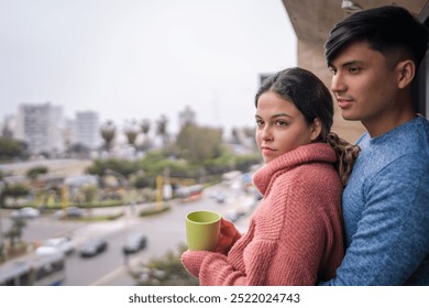 Young couple is embracing on their balcony, enjoying a warm drink and looking out at the city view - Powered by Shutterstock