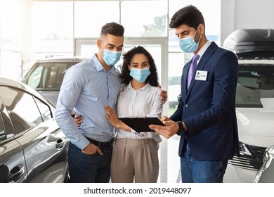 Young Couple Embracing While Having Conversation With Salesman, Looking At Papers In His Hands, Wearing Medical Face Masks, Buying New Car While Coronavirus Pandemic, Auto Showroom Interior