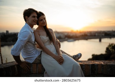 Young couple embracing while sitting on a wall, enjoying a beautiful golden hour sunset with a city river view