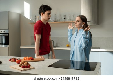 A young couple is engaged in a heated argument in a well-lit, contemporary kitchen setting. Tomato slices and a cucumber lay on a cutting board, suggesting dinner preparation was underway before the - Powered by Shutterstock