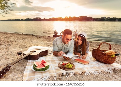 Young couple enjoying a picnic at the beach. Lying on the picnic blanket, reading books. White swans swimming and sunset over water in the background. Romance, dating and love concept.