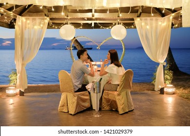 Young Couple Enjoying A Romantic Dinner By Candlelight, Outdoor.  Romantic Meal On The Beach With Lanterns, Chairs And Decorations, Sky, Sea And Beach In The Background. Loving Couple Eating Dinner.