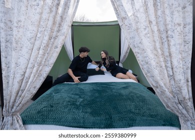 Young couple enjoying a romantic glamping getaway, relaxing and talking on a comfortable canopy bed inside a luxurious tent, surrounded by elegant curtains - Powered by Shutterstock - Get 15% off with code: PIKWIZARD15