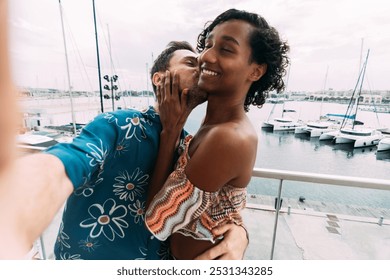 Young couple is enjoying their vacation by taking a selfie while kissing on a balcony overlooking a marina full of boats. Transgender woman. - Powered by Shutterstock