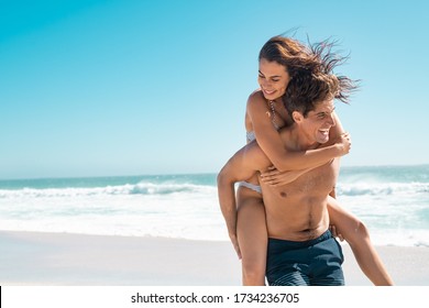 Young couple enjoying vacation at beach, copy space. Portrait of mid smiling man carrying girlfriend on his back along the sea shore. Boyfriend giving piggyback ride to happy woman having fun together - Powered by Shutterstock