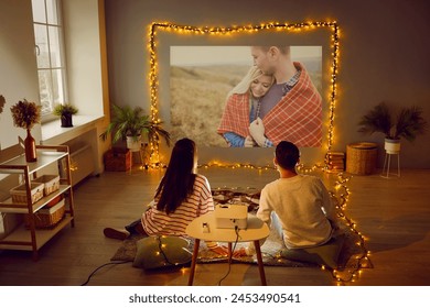 Young couple enjoying Valentine's Day, having romantic date at home, having good time together, watching movie on modern projector in cozy living room interior with beautiful fairy lights. Back view - Powered by Shutterstock