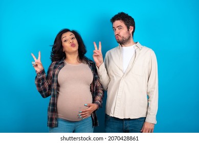 Young Couple Expecting A Baby Standing Against Blue Background Makes Peace Gesture Keeps Lips Folded Shows V Sign. Body Language Concept