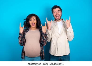 Young Couple Expecting A Baby Standing Against Blue Background  Makes Rock N Roll Sign Looks Self Confident And Cheerful Enjoys Cool Music At Party. Body Language Concept.