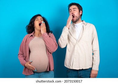 Young Couple Expecting A Baby Standing Against Blue Background Being Tired And Yawning After Spending All Day At Work.