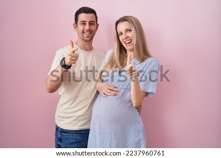 Young couple expecting a baby standing over pink background pointing fingers to camera with happy and funny face. good energy and vibes. 