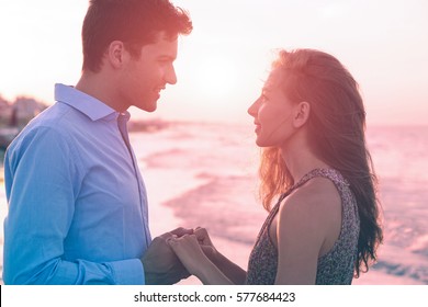 Young couple face to face holding hands each other standing on the beach shore at sunset - Happy lovers profile by the ocean gazing into eyes with passionate feeling - Bluish filter with sun halo 