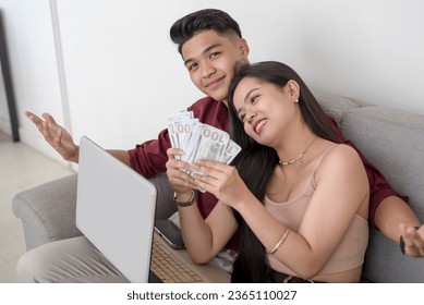 A young couple flaunting the earnings from their remote work via the laptop. A young wife holding multiple dollar bills and her husband bragging about their wealth.. - Shutterstock ID 2365110027