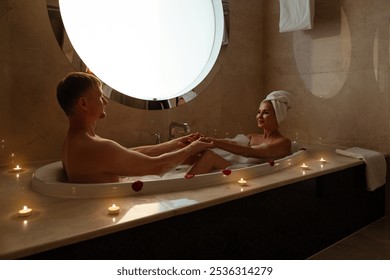 Young couple in an foam bath. Husband and wife in love Look at each other and hold hands sitting in bath. Newlyweds feast on bath. Couple in bathroom is relax on vacation in Jacuzzi. Candles, romance. - Powered by Shutterstock