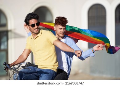 Young Couple Of Friends Holding A Gay Pride Flag While Riding A Bike Outdoors. LGBT, Relationship And Equal Rights Concept.