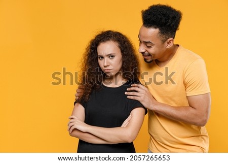 Young couple friends together family african frowning resentful woman man 20s wear black yellow t-shirt hugging offended girlfriend reconciling apologizes isolated on orange background studio portrait