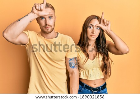 Young couple of girlfriend and boyfriend hugging and standing together making fun of people with fingers on forehead doing loser gesture mocking and insulting. 