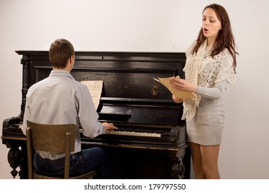 Young Couple Giving A Classical Duet With An Attractive Stylish Young Woman Singing From A Music Score Accompanied By A Young Man On A Piano