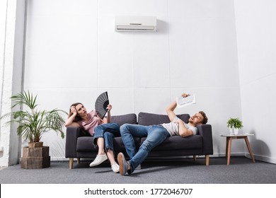 young couple with hand fan and newspaper suffering from heat at home with broken air conditioner