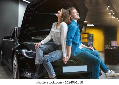 Young Couple Handsome Husband And Beautiful Wife Sitting On Open Car Bonnet In Electric Car Dealership Center, Eco Friendly Concept.