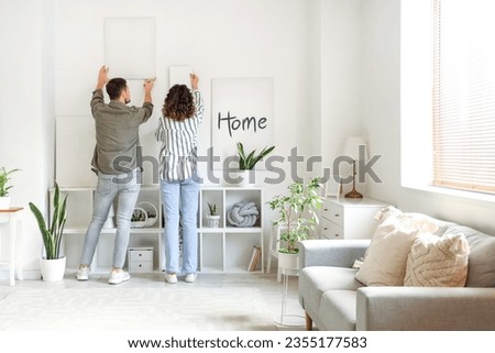 Young couple hanging paintings on light wall at home, back view