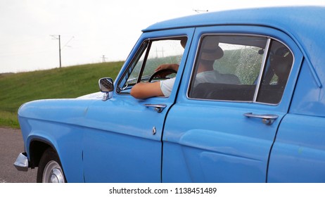 Young Couple In Hats Driving At Blue Retro Car After Rain During Summer Travel. Unrecognizable Pair Moving In Old Vintage Vehicle On Highway. Slow Motion Close Up Side View.