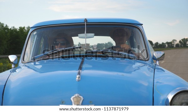 Young couple in hats riding in vintage car at summer\
travel. Man and woman sitting at the front seat of old retro car\
and talking during ride. Concept of road trip at holidays . Slow\
motion Close up.