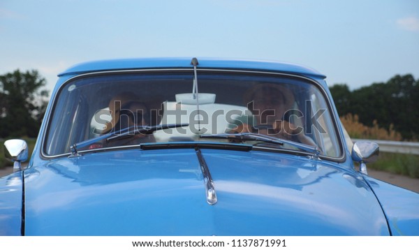 Young couple in hats riding in vintage car at summer\
travel. Man and woman sitting at the front seat of old retro car\
and talking during ride. Concept of road trip at holidays . Slow\
motion Close up.