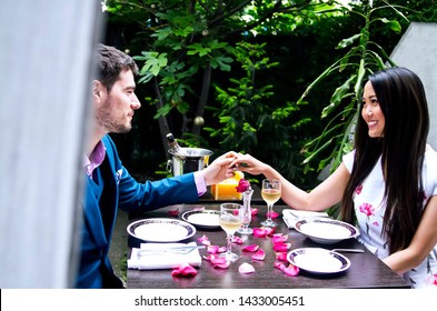 Young couple have romantic lunch in building garden.Chinese woman and Caucasian man holding hands above table with red rose petals and arranged plates with white wine in glasses