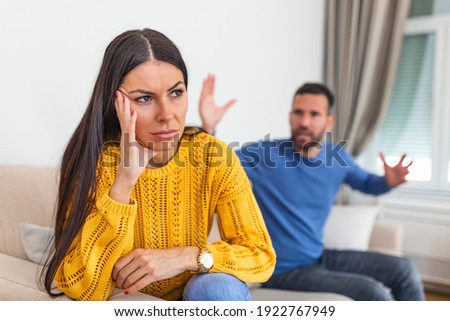 Young couple having an argument while sitting on their couch at home. Tired frustrated wife ignoring angry husband arguing blaming upset woman of problems, jealous man shouting at sad girlfriend