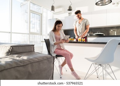 Young Couple Having Breakfast, Asian Woman Using Tablet Computer Hispanic Man Cooking Food Kitchen Modern Apartment Interior - Powered by Shutterstock