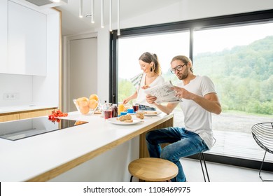 Young couple having breakfast cooking some food and reading newspaper at the kitchen of the modern country house with green area outdoors - Powered by Shutterstock