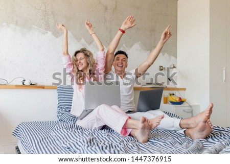 young couple having fun on bed in morning holding hands up, man and woman with laptop, online freelancer job, smiling happy family together, bedroom pajamas