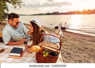 Young couple having a picnic at the beach. Lying on the picnic blanket, drinking red wine. White swans swimming and sunset over water in the background. Romance, dating and love concept. - Powered by Shutterstock