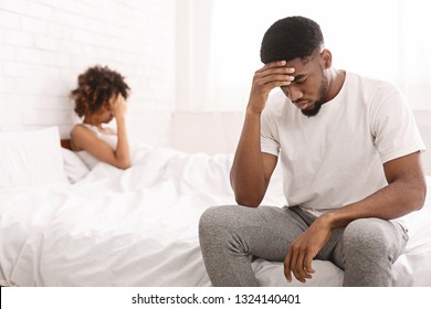 Young couple having problems. Desperate black guy sitting on bed and thinking, his girlfriend on background, free space