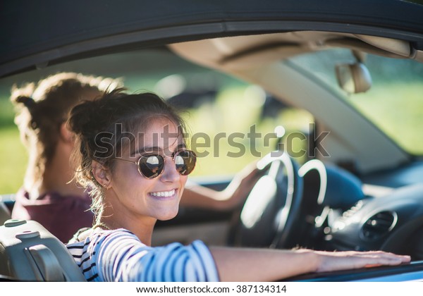 Young couple in his car, happy to drive on a\
country road, focus on the woman. Shot with flare. There are some\
cows at the background