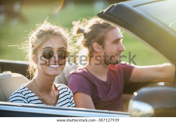 Young couple in his convertible car, happy\
to drive on a country road, focus on the woman. Shot with\
flare.There are some blurred cows at the\
background