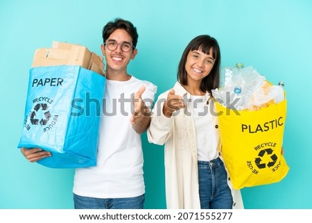 Young couple holding a bag full of plastic and paper to recycle isolated on blue background shaking hands for closing a good deal